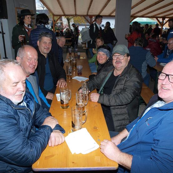 Eine Gruppe von Männern sitzt an einem langen Tisch in einem Biergarten im Freien, lächelt und hält Gläser mit Bier. Hinter ihnen sitzen weitere Personen, einige stehen und andere gehen herum.