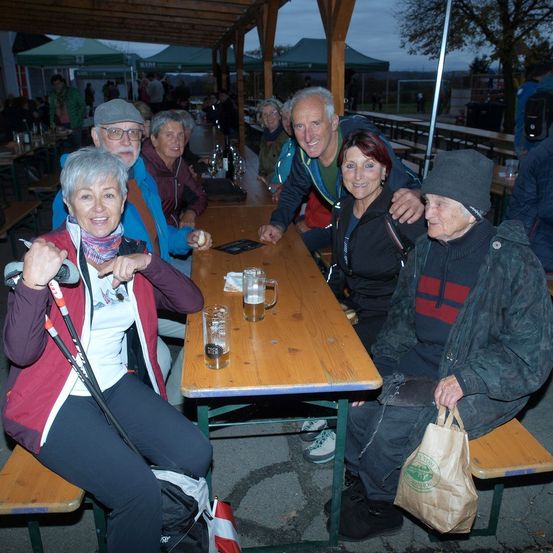 Mehrere Erwachsene, wahrscheinlich eine Familie, sitzen an einem Tisch im Freien, möglicherweise in einem Restaurant. Eine Frau hält eine Skistöcke. Ein Mann neben ihr lächelt, während ein anderer Mann und eine Frau Händchen halten. Vor ihnen steht ein Tisch mit zwei Gläsern und einem Krug. Im Hintergrund sitzen weitere Personen an Tischen, und Zelte sind sichtbar.