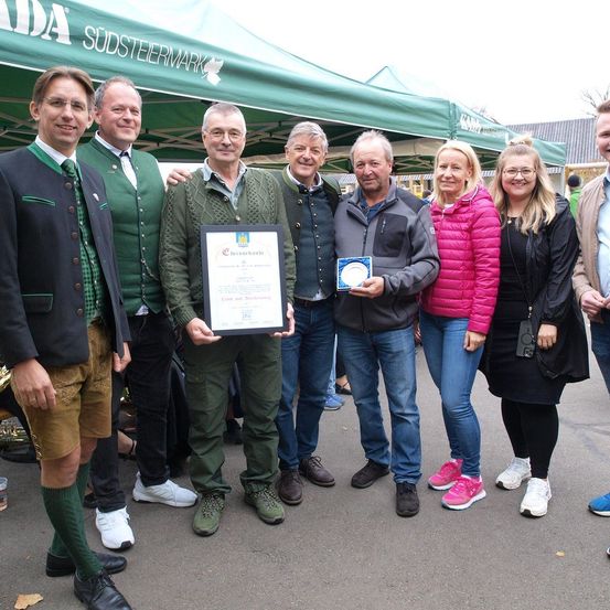 Sechs Personen stehen in einer Gruppe unter einem Zelt. Ein Mann hält ein gerahmtes Zertifikat. Ein Mann neben ihm trägt einen grünen Pullover. Die anderen sind lässig gekleidet. Hinter ihnen ist ein grünes Zelt zu sehen.