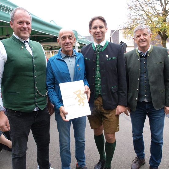 Vier Männer stehen lächelnd zusammen. Einer hält ein Zertifikat. Sie tragen traditionelle Kleidung. Ein grünes Zelt und ein Baum sind im Hintergrund.