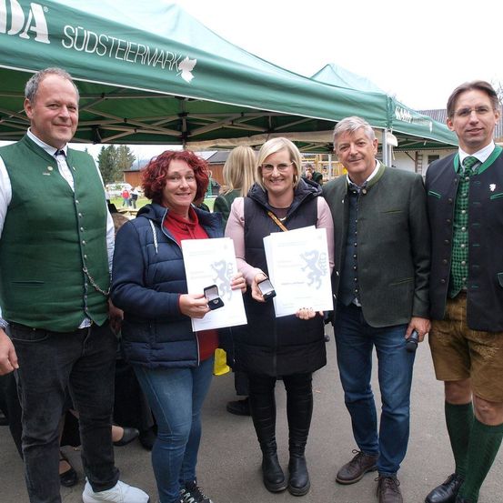 Fünf Personen, darunter zwei Frauen, stehen unter einem grünen Zelt, halten Urkunden und lächeln für die Kamera. Hinter ihnen sind verschwommene Menschen und Bäume zu sehen.