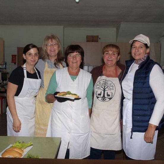 Fünf Frauen in Schürzen stehen lächelnd in einer Küche. Eine hält einen Teller mit Sandwiches. Hinter ihnen sind Schränke und ein Feuerlöscher zu sehen. Ein Tisch mit Sandwiches steht im Vordergrund.