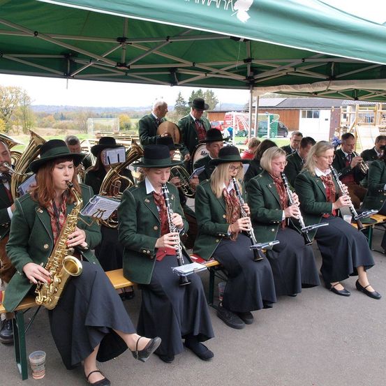 Eine Bläserkapelle aus Frauen und Männern sitzt unter einem grünen Zelt. Sie sind in grünen Uniformen und Hüten gekleidet. Sie spielen Blasinstrumente. Im Hintergrund sind ein Sportplatz, ein Spielplatz und einige Gebäude zu sehen.