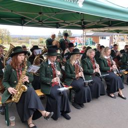 Eine Bläserkapelle aus Frauen und Männern sitzt unter einem grünen Zelt. Sie sind in grünen Uniformen und Hüten gekleidet. Sie spielen Blasinstrumente. Im Hintergrund sind ein Sportplatz, ein Spielplatz und einige Gebäude zu sehen.