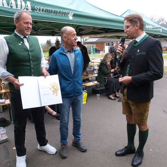 Drei Männer stehen unter einem Zelt. Ein Mann hält ein Papier, ein anderer spricht in ein Mikrofon und der dritte schaut sie an. Hinter ihnen sitzen mehrere Menschen auf Bänken.
