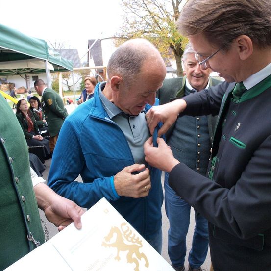Ein Mann in einer blauen Jacke wird von einem anderen Mann in einem Anzug mit einer Medaille ausgezeichnet. Andere Leute stehen um sie herum. Ein grünes Zelt ist hinter ihnen.