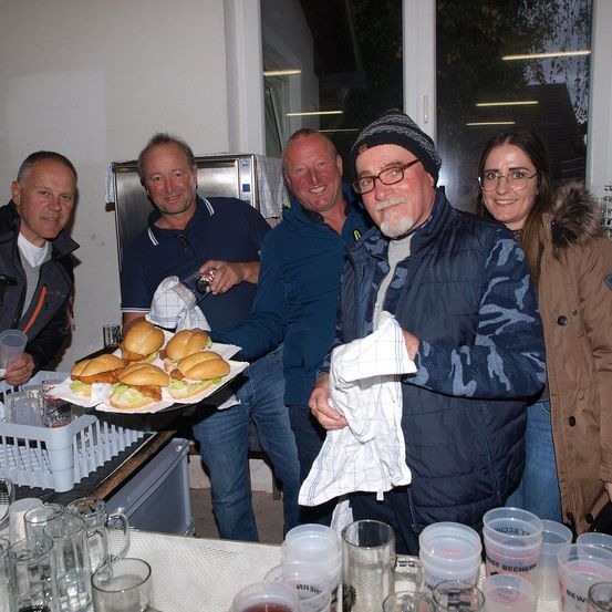 Fünf Menschen lächeln und posieren für ein Foto in einer Küche. Ein Mann hält einen Tablett mit Sandwiches, andere halten Becher und Gläser. Ein Kühlschrank und ein Korb sind auf dem Tresen.