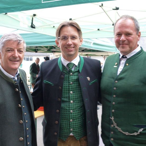 Drei Männer in traditioneller grüner Kleidung lächeln unter einem grünen Zelt. Der Mann auf der linken Seite trägt einen grünen Mantel mit Knöpfen. Der Mann in der Mitte trägt ein grünes und schwarzes Outfit mit einer grünen Krawatte. Der Mann auf der rechten Seite trägt eine grüne Weste mit einer silbernen Kette.