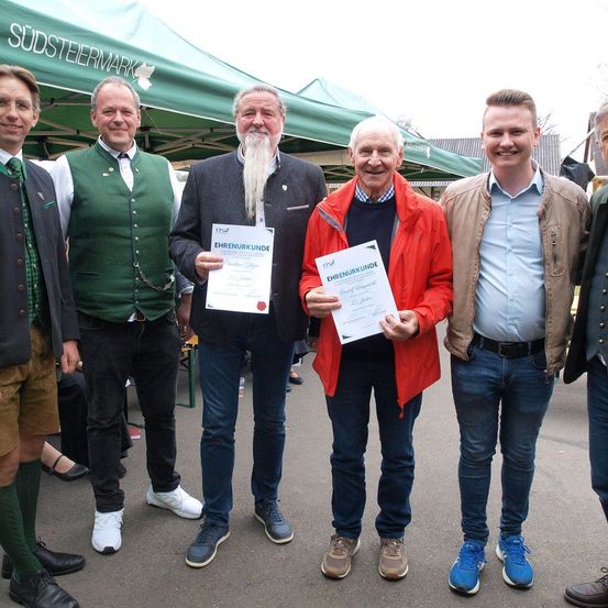 Sechs Männer stehen in einer Reihe, zwei halten Urkunden. Der Mann im roten Mantel hält eine Urkunde mit der Aufschrift 'Ehrenurkunde'. Hinter ihnen ist ein grünes Zelt mit dem Text 'Sudsteiermark' zu sehen.