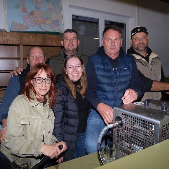 Fünf Menschen stehen drinnen und lächeln. Zwei Männer und zwei Frauen sind vorne, und ein Mann steht hinter ihnen. Sie stehen um eine Maschine mit einem Schlauch herum. Eine Frau hält ein Glas. Hinter ihnen ist ein Regal mit einer Karte und einem Fenster.