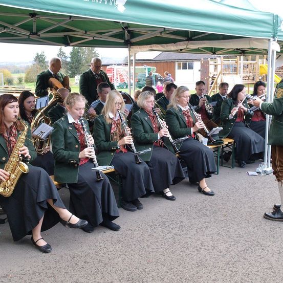 Eine Gruppe von Musikern in grünen Uniformen sitzt unter einem grünen Zelt und spielt verschiedene Instrumente. Ein Dirigent steht vor ihnen.