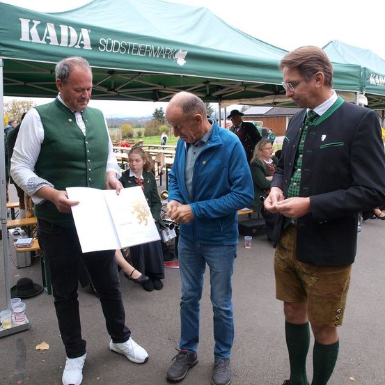 Drei Männer versammeln sich unter einem grünen Zelt mit der Aufschrift KADA. Einer hält ein offenes Buch, während die anderen etwas betrachten. Eine Frau in einer Schuluniform sitzt in der Nähe. Die Umgebung deutet auf eine ungezwungene Veranstaltung im Freien hin.