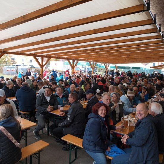 Eine Menschenmenge versammelt sich in einem überdachten Zelt mit Holztischen und -bänken. Einige sitzen, andere stehen. Viele tragen Hüte, Brillen und Jacken. Sie lächeln und unterhalten sich. Einige halten Gläser mit Bier in der Hand. Das Zelt hat einen Holzrahmen und ein weißes Dach.