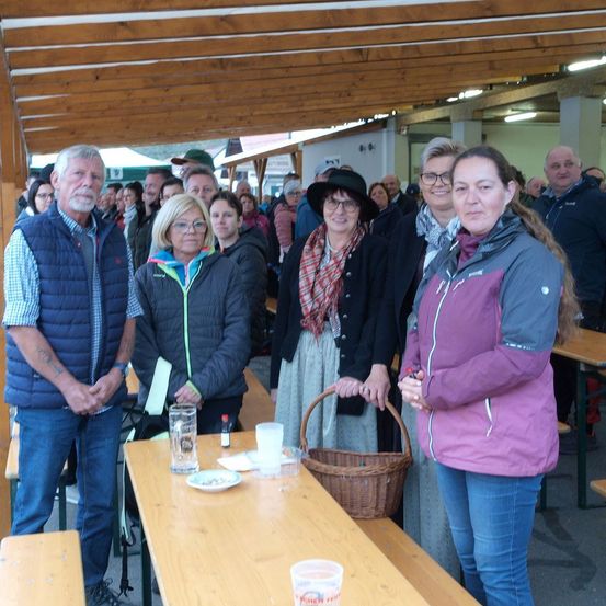 Eine Gruppe von Menschen steht unter einem hölzernen Pavillon, mit einem Tisch vor ihnen. Eine Frau in einer lila Jacke hält einen Korb. Hinter ihnen steht ein Mann mit einer blauen Weste. Die Umgebung scheint eine Versammlung oder ein Ereignis zu sein.