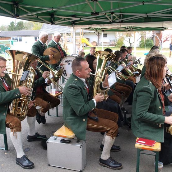 Ein Blasorchester spielt unter einem grünen Zelt, die Musiker tragen grüne Uniformen und Lederschuhe. Sie spielen Instrumente wie Tubas und Trommeln, während andere Noten lesen. Das Publikum sitzt in der Nähe.
