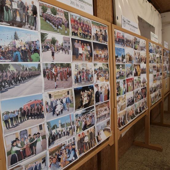 Eine Reihe von Fotografien ist an einer Holztafel befestigt und zeigt verschiedene Veranstaltungen und Zusammenkünfte. Die Bilder zeigen Menschen in traditioneller Kleidung, Paraden, gesellige Zusammenkünfte und Gemeinschaftsveranstaltungen.