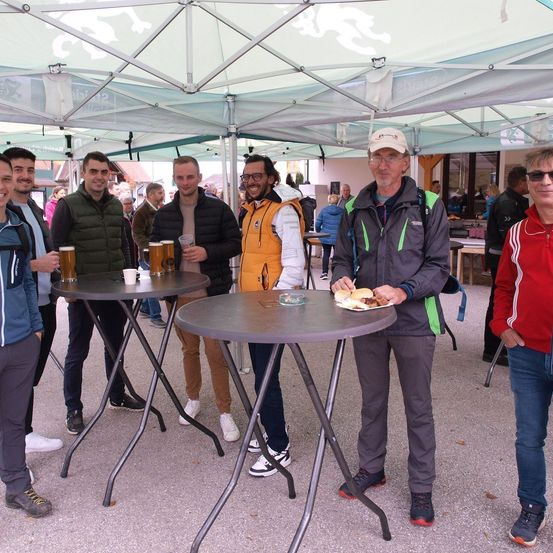 Eine Gruppe von Männern steht unter einem Zelt, einige halten Getränke, andere essen. Ein Mann in einer Jacke hält einen Teller mit Essen. Der Bereich ist im Freien mit Tischen und einem Baldachin.