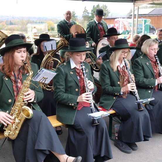 Eine Gruppe von Menschen sitzt auf einer Bank und spielt Musikinstrumente, wahrscheinlich im Freien. Sie tragen Uniformen und Hüte. Einige spielen Saxophon, Trompete und Klarinette. Hinter ihnen stehen und sitzen weitere Menschen auf einer Bank.