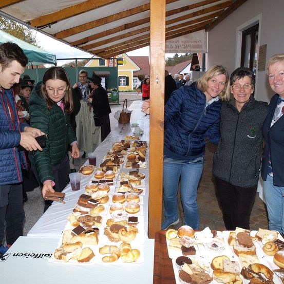 Eine Gruppe von Menschen versammelt sich um einen Tisch voller verschiedener Gebäcke vor einem Gebäude. Sie lächeln und scheinen das Essen zu genießen. Ein Mann in einem blauen Mantel macht ein Foto von den Gebäcken.