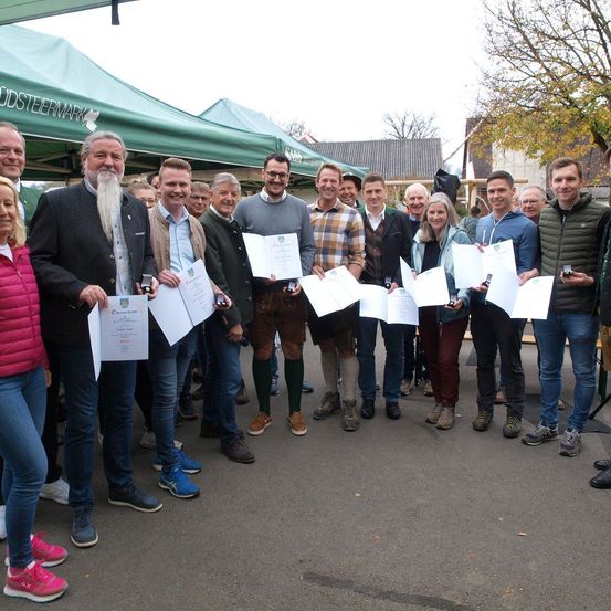 Gruppe von Menschen in traditioneller Kleidung, lächelnd und Urkunden haltend, stehen im Kreis mit Zelten und Gebäuden im Hintergrund.