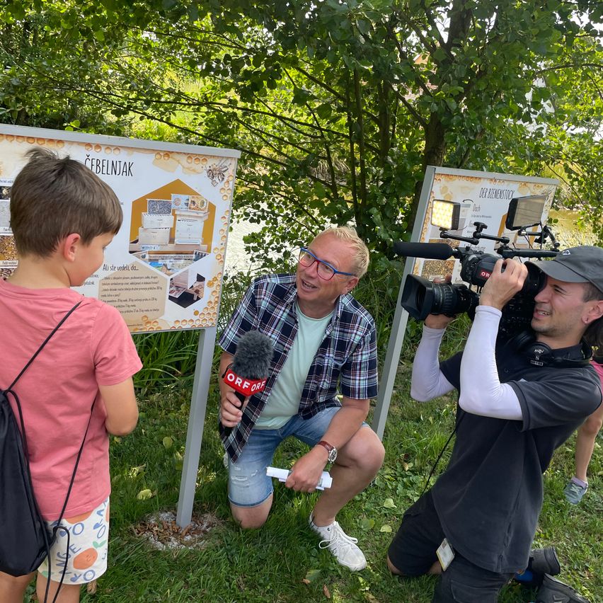 Ein Mann interviewt einen Jungen vor einem Informationsschild. Ein anderer Mann filmt mit einer Kamera. Der Junge trägt einen Rucksack. Bäume sind im Hintergrund.