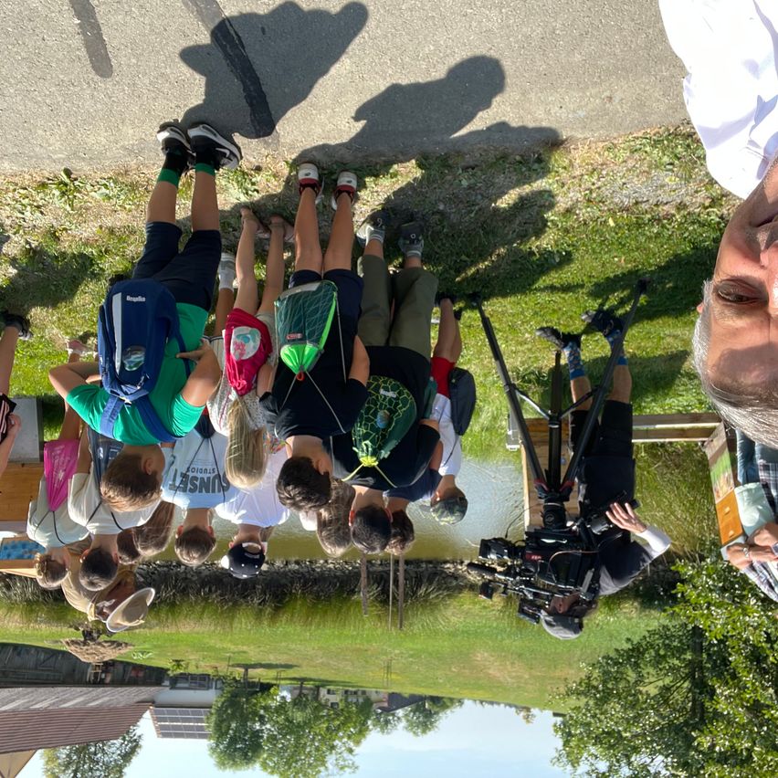 Eine Gruppe von Menschen steht auf einem Rasen und lächelt für die Kamera. Sie tragen Rucksäcke, und einige von ihnen tragen Turnschuhe. Eine Person hält eine Kamera. Dahinter befindet sich ein Gewässer, und auf beiden Seiten stehen einige Bäume.