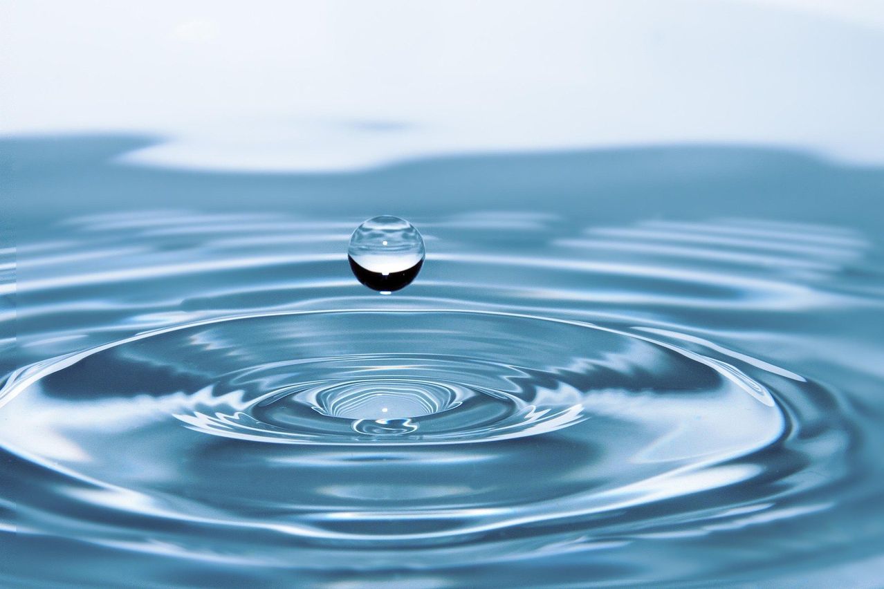 Ein Wassertropfen schwebt über der Wasseroberfläche und erzeugt Wellen und Reflexionen. Das Wasser erscheint ruhig und klar.