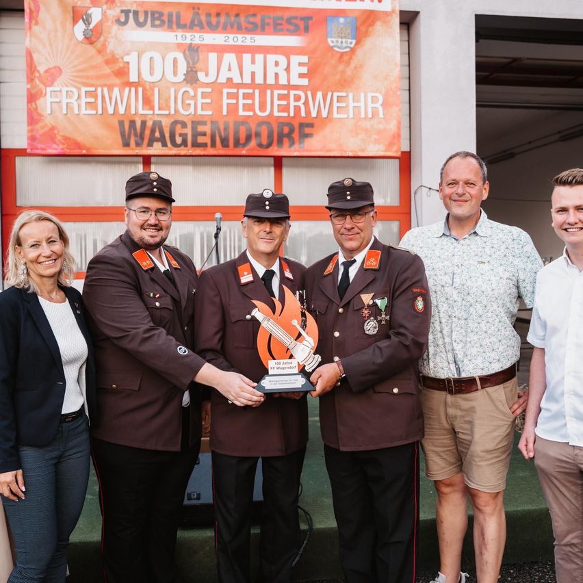 Eine Gruppe von fünf Personen steht vor einem Banner mit der Aufschrift '100 Jahre Freiwillige Feuerwehr Wagendorf'. Die beiden Männer in der Mitte halten einen Pokal mit Flammendesign. Alle sind in Uniformen gekleidet.