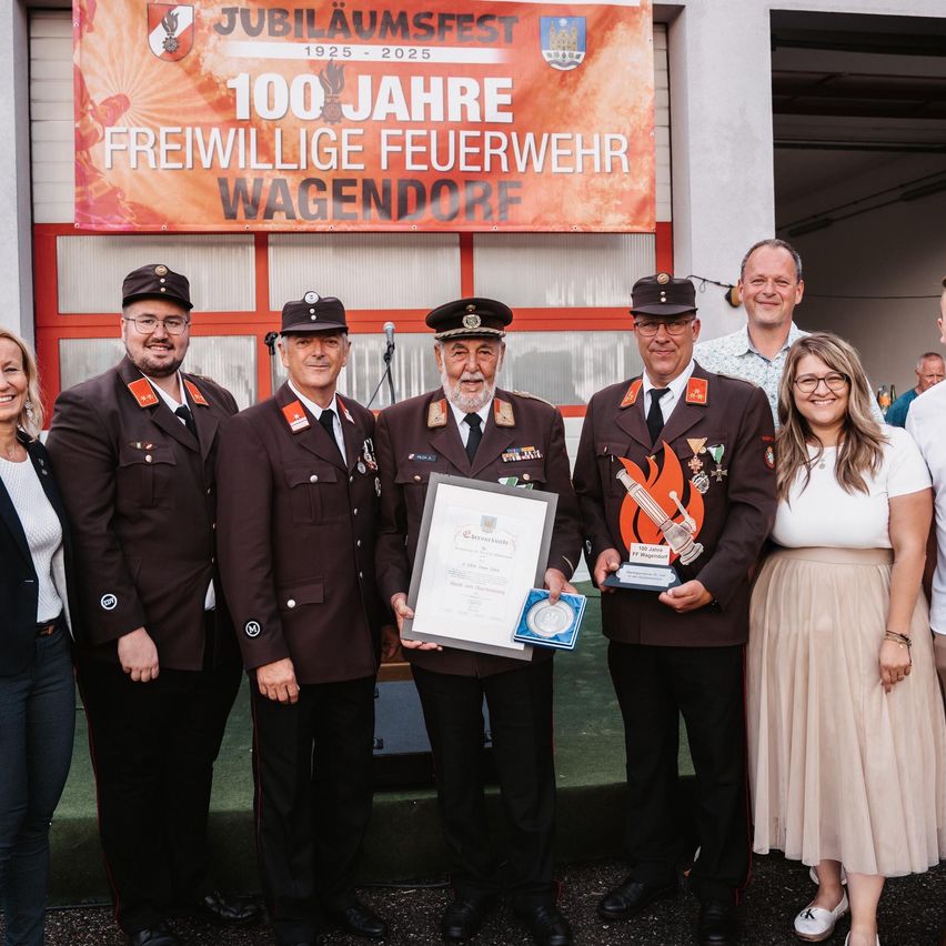 Eine Gruppe von Menschen in braunen Uniformen posiert für ein Foto. Ein Mann in der Mitte hält eine Plakette. Ein Banner hinter ihnen lautet '100 Jahre Freiwillige Feuerwehr Wagendorf.'