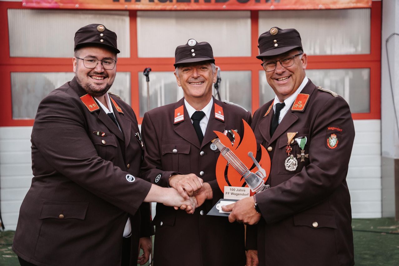 Drei Männer in Militäruniformen, lächelnd, halten Hände um eine Trophäe mit der Aufschrift '100 Jahre FF Wagendorf', stehen vor einem Gebäude mit roten Akzenten.