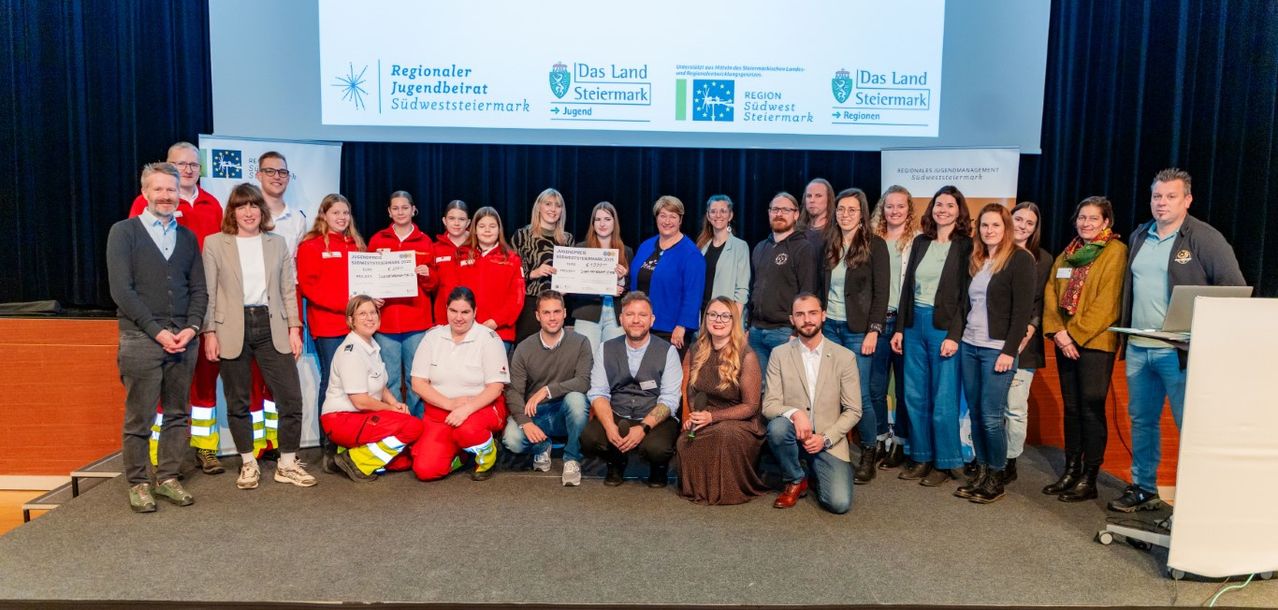 Eine Gruppe von Menschen in Uniformen und Anzügen posiert für ein Foto auf der Bühne mit einem Bildschirm im Hintergrund.