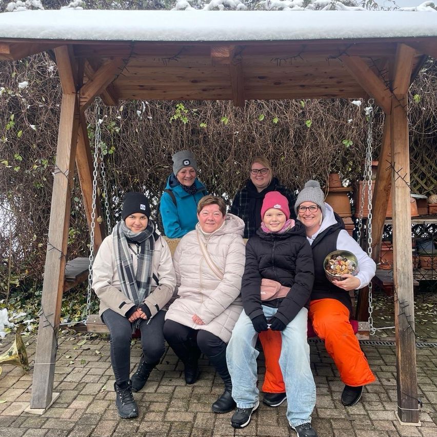 Eine Gruppe von sechs Personen, die warm gekleidet für kaltes Wetter sind, posiert in einem schneebedeckten Holzpavillon. Sie sitzen auf einer Schaukel, wobei eine Person eine Schüssel mit Essen hält.