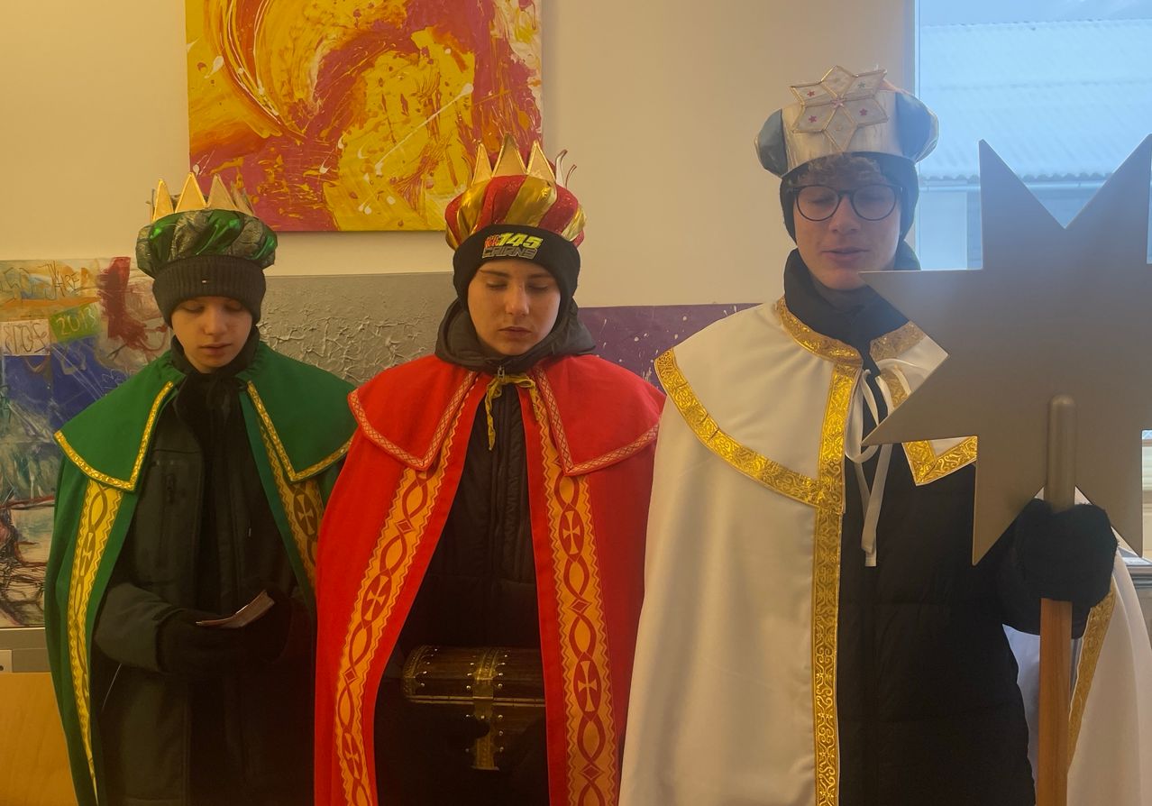 Drei Menschen in aufwendigen Kostümen stehen zusammen, einer liest aus einem Buch, einer hat ein Buch in der Hand und ein dritter trägt eine Krone. Sie stehen vor einem farbenfrohen Gemälde.