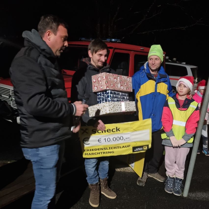 Drei Männer und ein Kind stehen draußen mit einem roten Fahrzeug im Hintergrund. Sie halten eine Geschenkbox und einen Spendenbeleg. Das Kind trägt einen Weihnachtsmannhut.