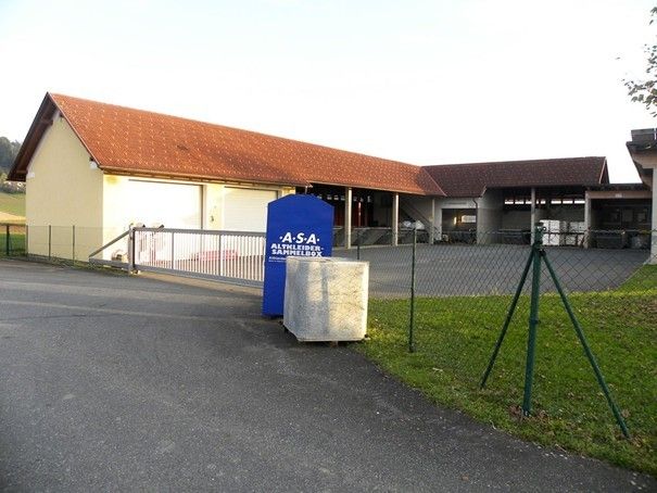 Ein Gebäude mit einem braunen Dach und einem blauen Schild mit der Aufschrift 'A.S.A. Sammelbox'. Das Gebäude hat eine Garage und einen Zaun. Auf der Einfahrt steht ein großer Felsblock.