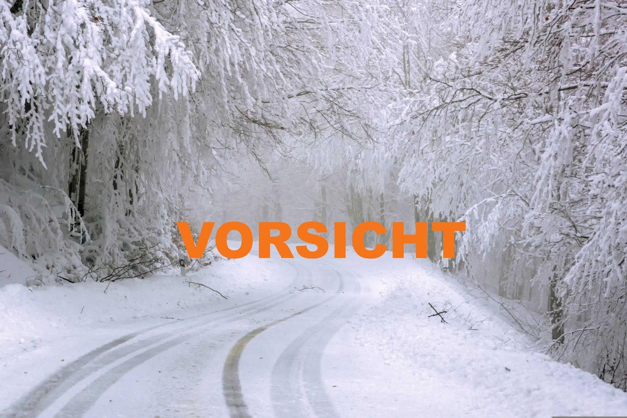 Verschneiter Weg mit schneebedeckten Bäumen. Der orange Text 'VORSICHT' warnt Fahrer. Die Straße ist rutschig.