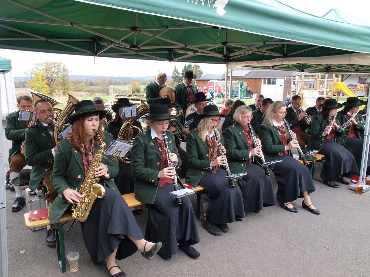 Eine Gruppe von Musikern in grünen Uniformen und Hüten sitzt auf Bänken unter einem grünen Zelt und spielt Instrumente. Dahinter befindet sich eine Rasenfläche mit Bäumen und Gebäuden.
