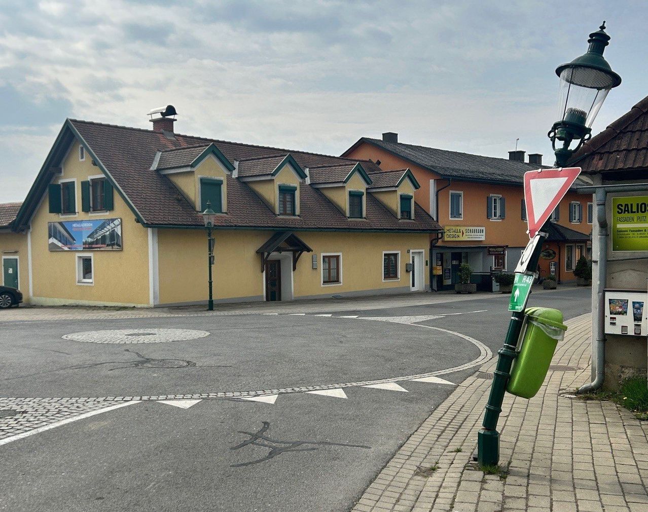 Eine ruhige Straße mit gelben Häusern, einer Straßenlaterne und einem dreieckigen Schild. Die Häuser haben braune Dächer und grüne Fensterläden. Ein Mülleimer ist auf dem Bürgersteig platziert.