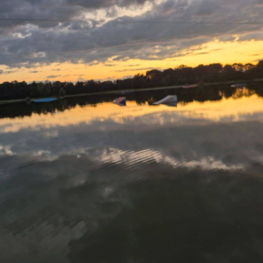 Ein ruhiger Sonnenuntergang über einem ruhigen See mit Reflexionen von Wolken und Bäumen. Zwei Objekte schwimmen im Wasser.