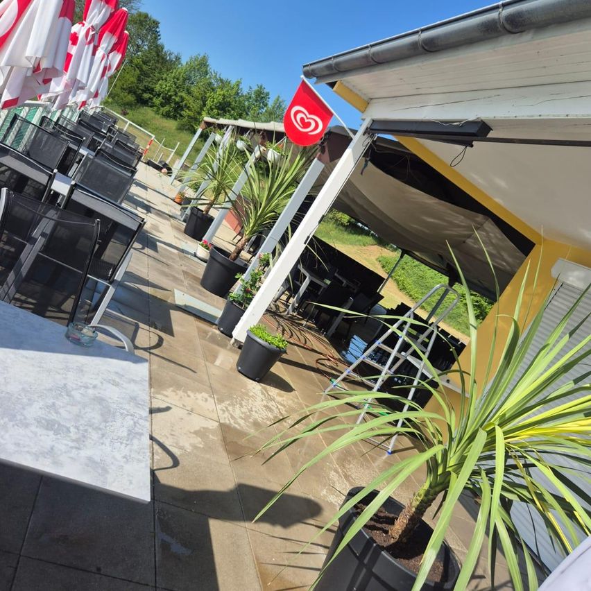 Eine Außengastronomie mit Reihen schwarzer Stühle unter roten und weißen Sonnenschirmen. Topfpflanzen und eine rote Flagge mit einem Herz sind auf der Seite. Ein Gebäude mit gelber Wand und einer Leiter befindet sich rechts.