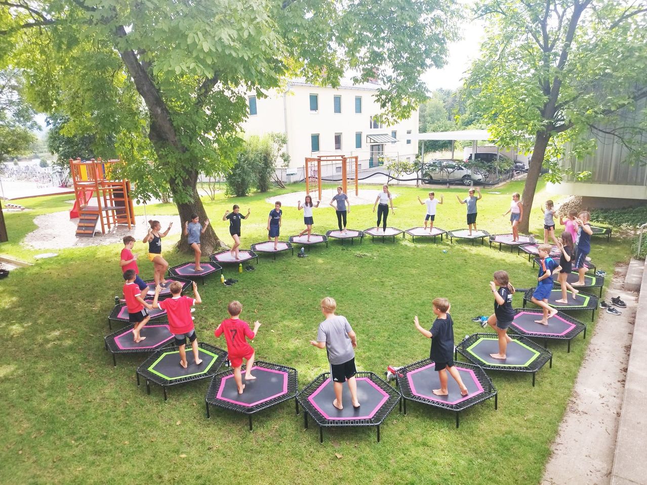 Kinder machen Übungen in einem Kreis auf Trampolinen in einem Park. Die Kinder tragen verschiedene farbige Hemden. Dahinter gibt es Bäume, ein Gebäude und Autos.