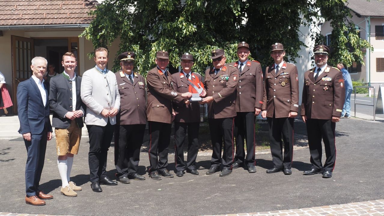 Sechs Männer in Uniformen stehen in einer Reihe, einer hält eine Trophäe und lächelt für ein Foto auf einer Straße.