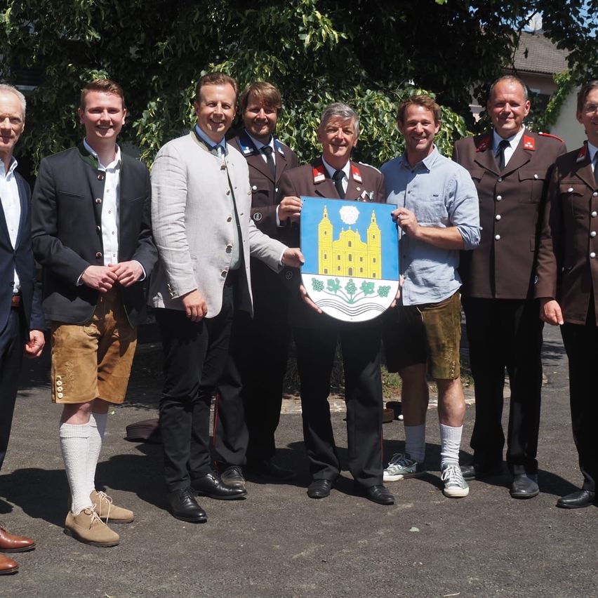 Eine Gruppe von Männern in verschiedenen Outfits posiert für ein Foto, einer hält ein blaues Schild mit Burgdesign. Sie stehen auf einem gepflasterten Bereich mit Bäumen im Hintergrund.