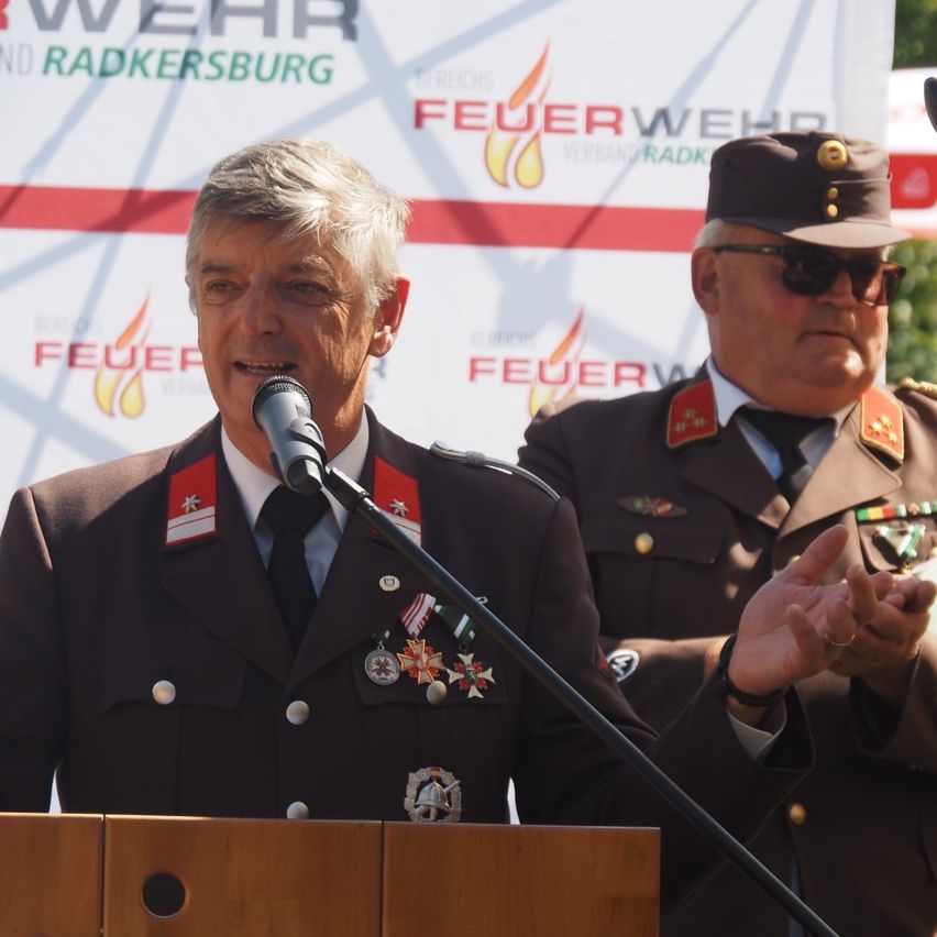 Ein Mann spricht in ein Mikrofon an einem Podium, während ein weiterer Mann in Uniform hinter ihm steht. Der Hintergrund zeigt Logos und Text im Zusammenhang mit der Feuerwehr.