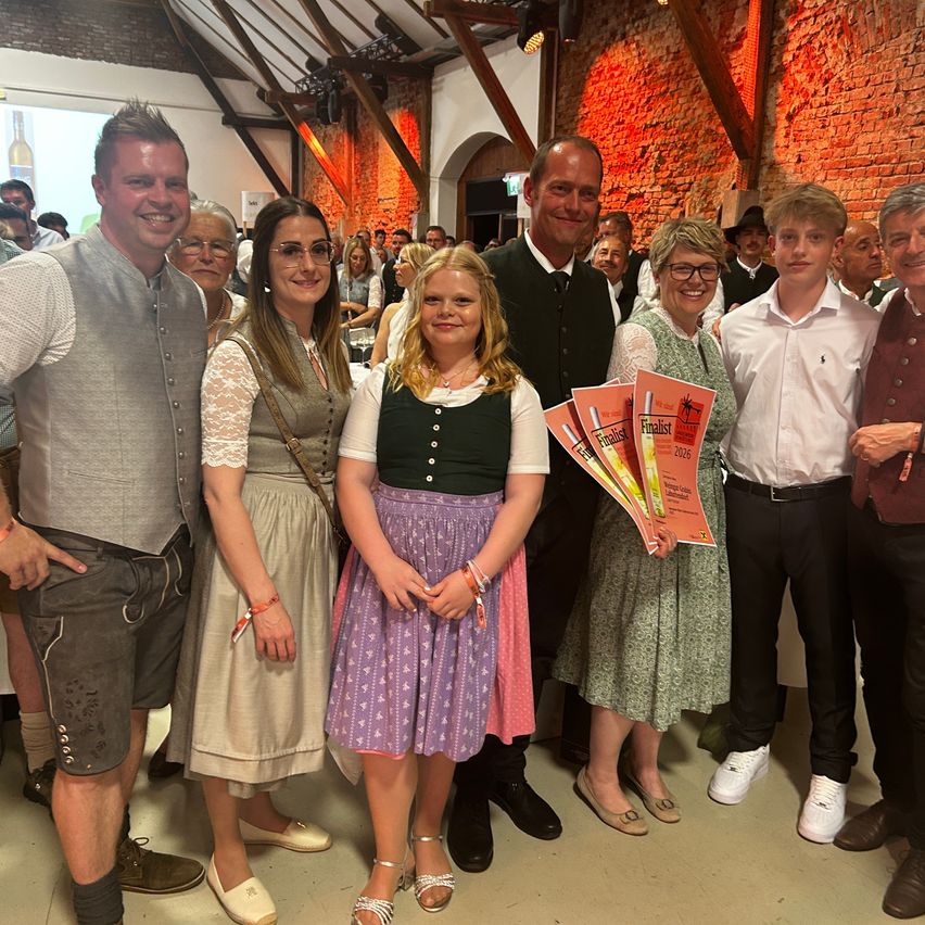 Eine Gruppe von Menschen in traditioneller Kleidung posiert für ein Foto und hält Zertifikate in der Hand, mit einer Ziegelwand im Hintergrund.