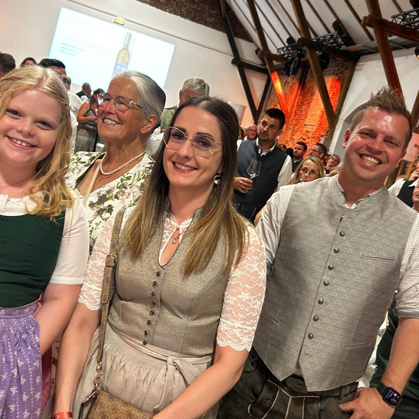 Drei Menschen lächeln und stehen eng beieinander und posieren für ein Foto. Dahinter versammelt sich eine große Menschenmenge im Innenraum. Eine Beamerleinwand zeigt eine Dia-Folie an.