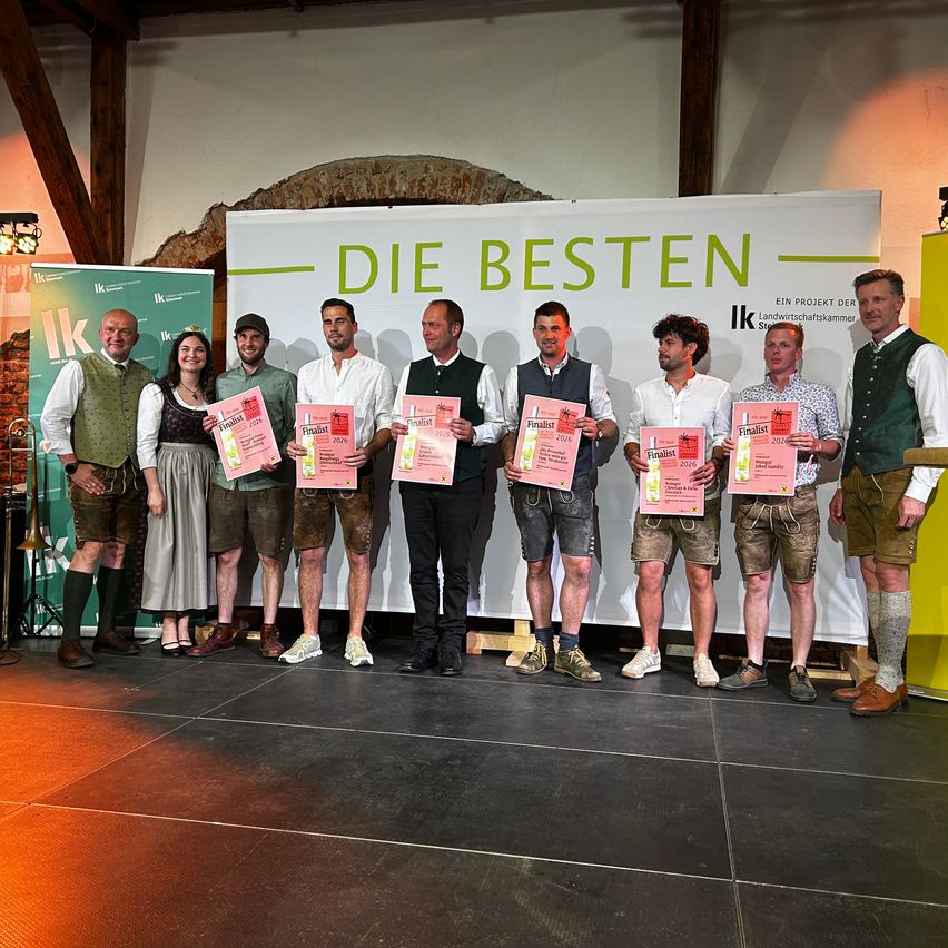 Gruppenfoto der Preisträger in traditioneller bayerischer Kleidung. Sie stehen auf der Bühne und halten Urkunden in der Hand vor einem Banner, das 'Die Besten' steht.