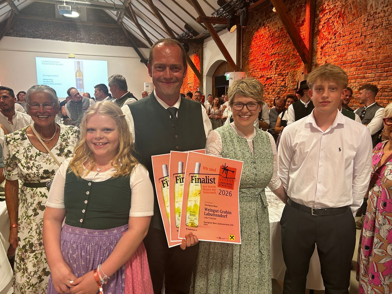 Eine vierköpfige Familie steht lächelnd vor einer Menschenmenge. Der Mann hält einen Finalisten-Poster für 2026. Eine Frau und ein Mädchen tragen traditionelle Kleidung.