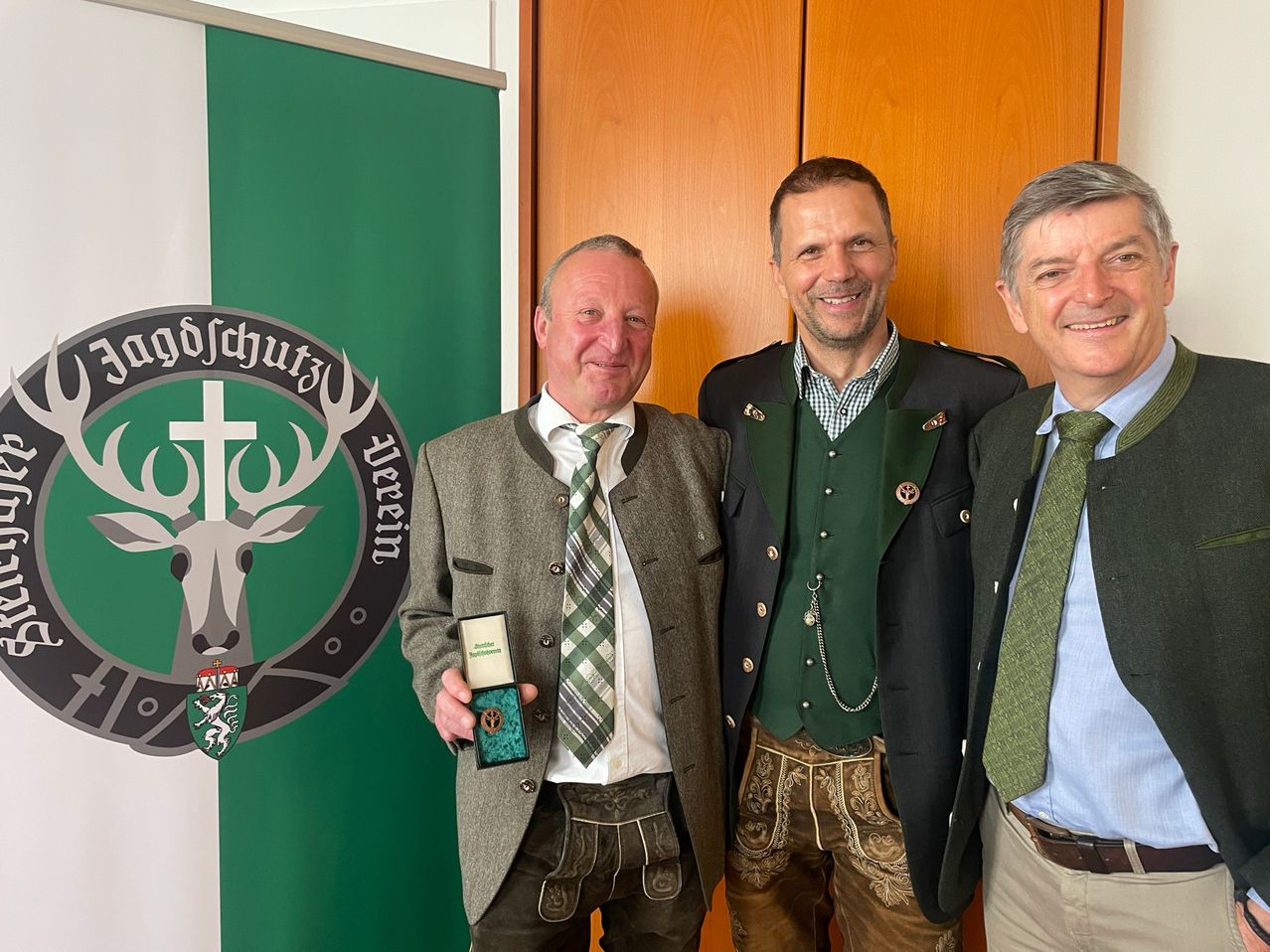 Drei Männer stehen lächelnd zusammen. Der Mann auf der linken Seite hält einen Preis. Sie stehen vor einer Holzwand und einem grünen Banner mit einem Logo.