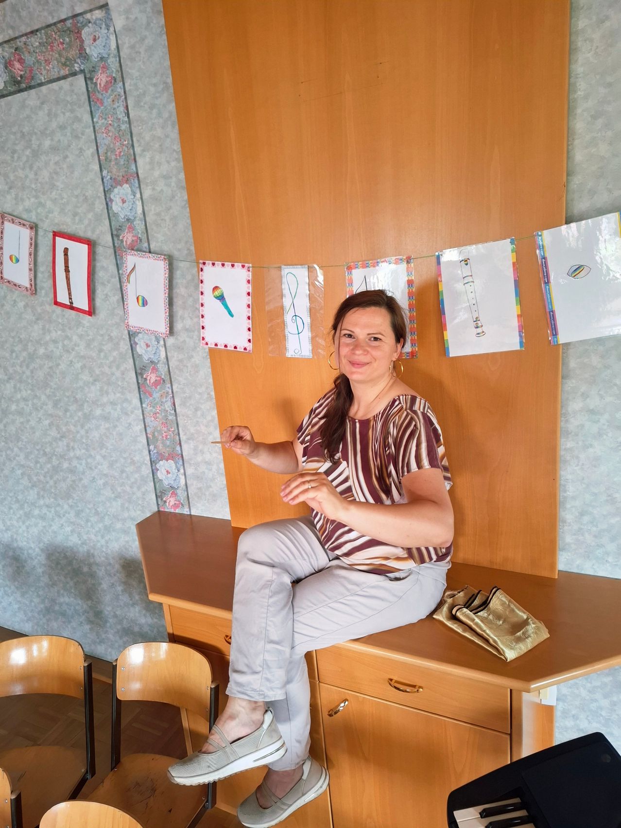 Eine Frau sitzt auf einem Holzschreibtisch und hält ein Musikinstrument. Hinter ihr zeigt eine Wand bunte Musiknoten und Instrumente.